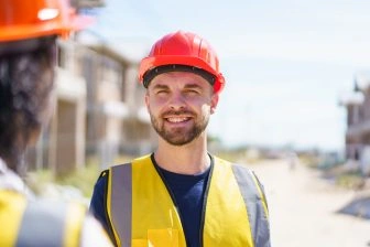 Ouvrier du bâtiment souriant portant casque et gilet de sécurité sur un chantier, illustrant les métiers du BTP et la prévention.